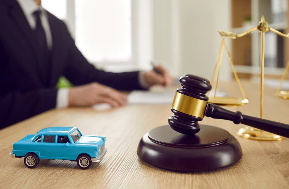 A small toy car sits on a table alongside a judge's gavel, set against the backdrop of a judge solemnly signing a legal document.