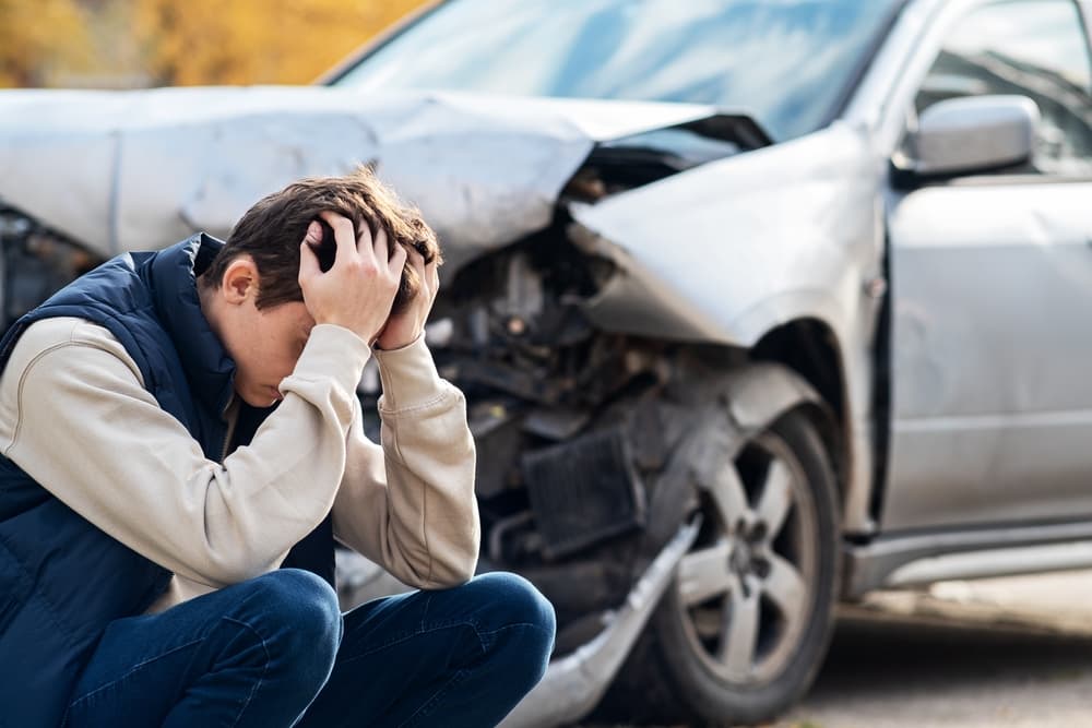 A man clutching his head in pain stands near a wrecked car, suddenly realizing the severity of his injury.