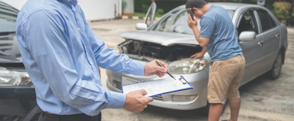 An insurance officer diligently fills out a claim report on their clipboard while overseeing the insurance claim process between a man and an insurance agent following a car crash.






