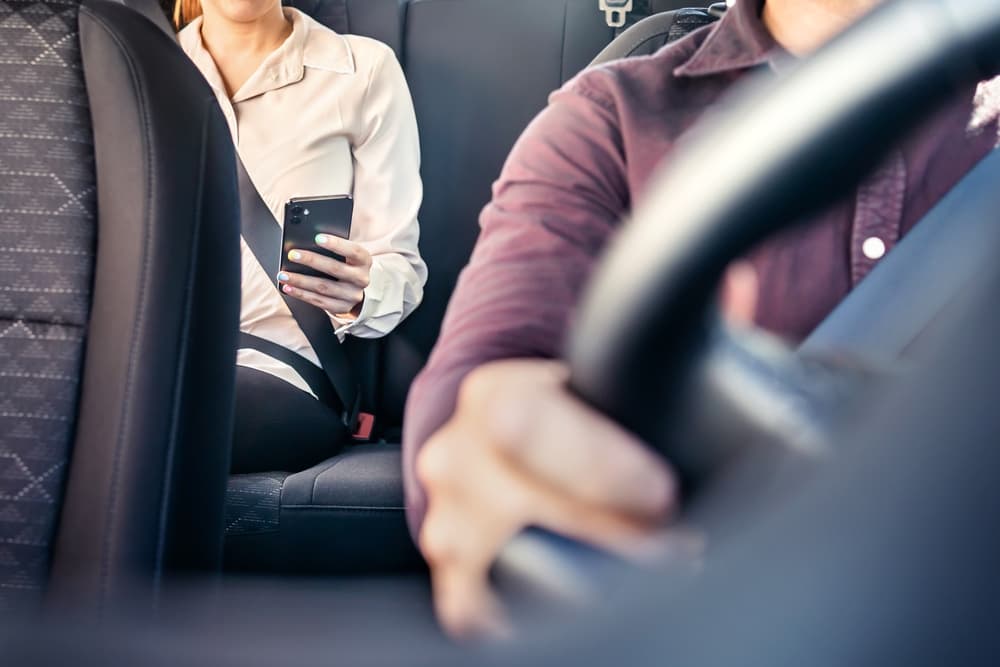 A professional businesswoman using her cellphone in the back seat of a taxi or rideshare car. The driver is in the front, and she is accessing a rideshare mobile app.