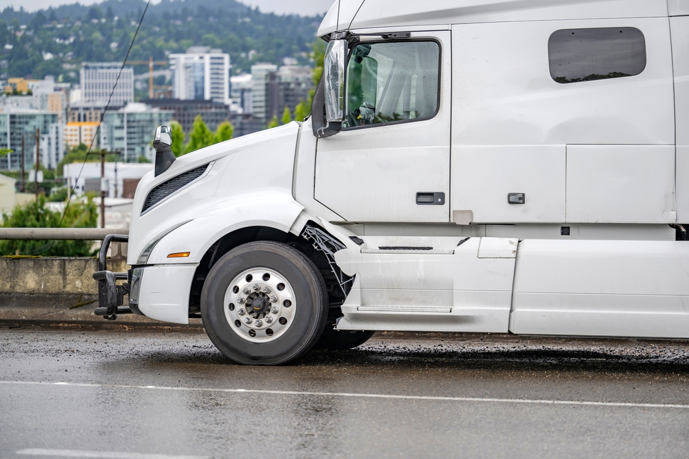 A big rig semi-truck with significant body damage from an accidental collision stands out of service on the highway shoulder, awaiting roadside assistance.