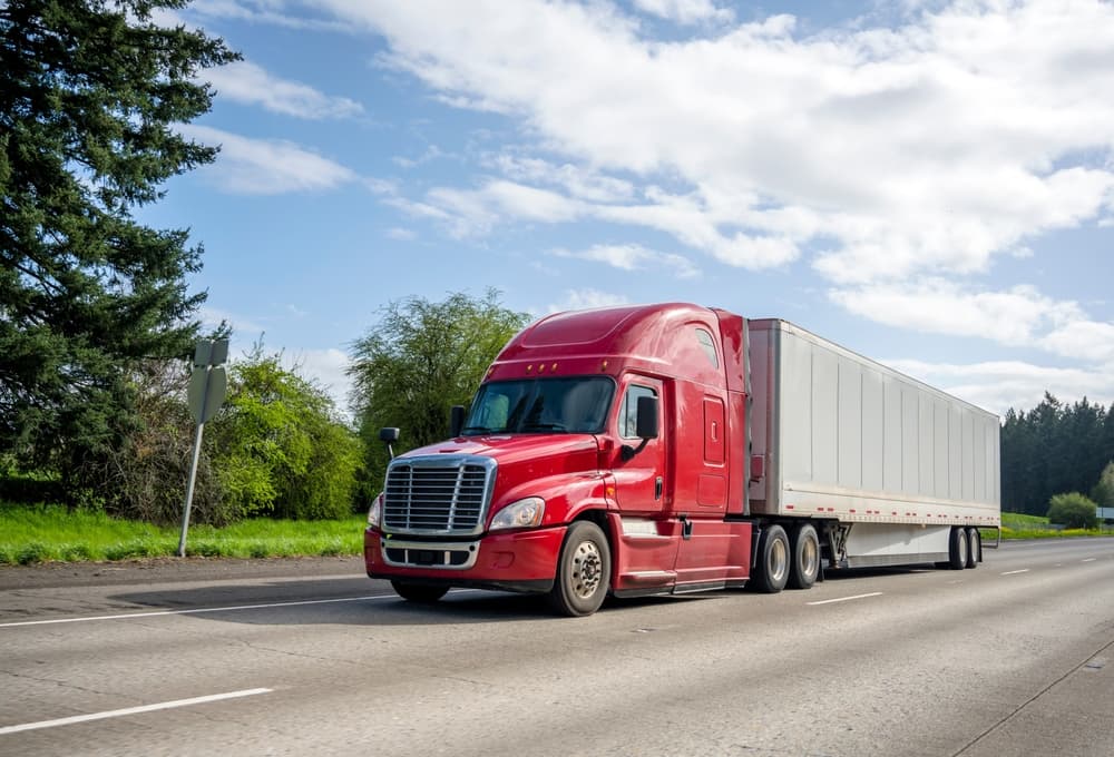 A red big rig, known for its reliability and popularity, transports commercial cargo in a dry van semi-trailer as it moves along a wide, straight highway.