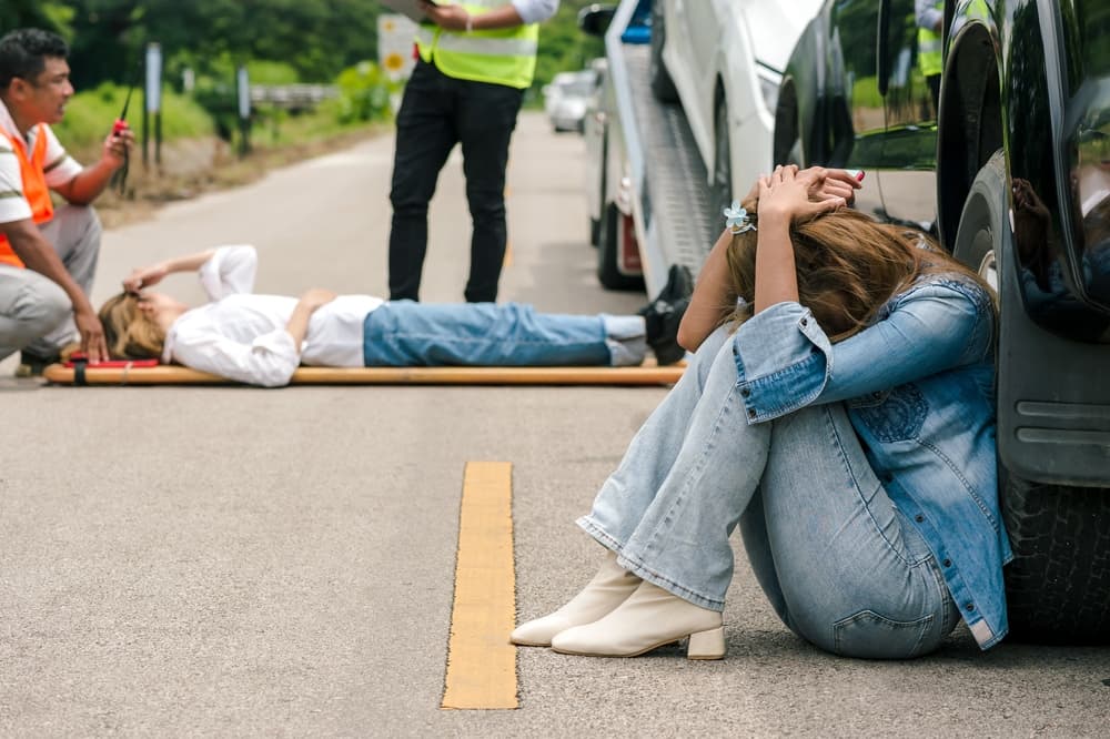 A drunk driver woman, in shock and distress, stands near the scene of a car accident involving a young female victim lying on a stretcher with a rescue team in the background.