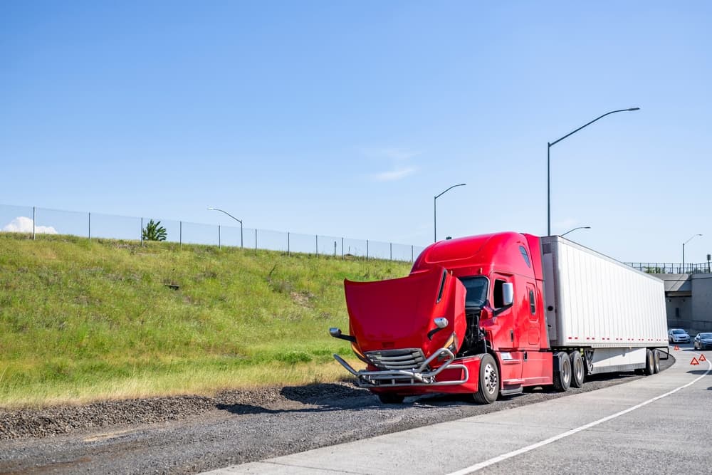 A broken-down red big rig semi truck with an open hood and dry van trailer stands out of service at a highway road entrance.