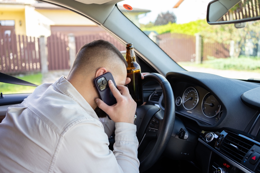 A man holds a mobile phone and a bottle of beer while driving, highlighting the dangers of alcohol use and distracted driving, increasing the risk of an accident.