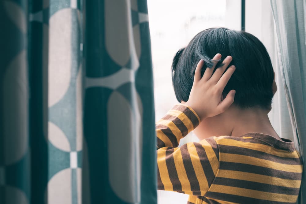 A young Asian boy stands by a window, crying and covering his ears, overwhelmed by fear, insecurity, anxiety, and stress. 