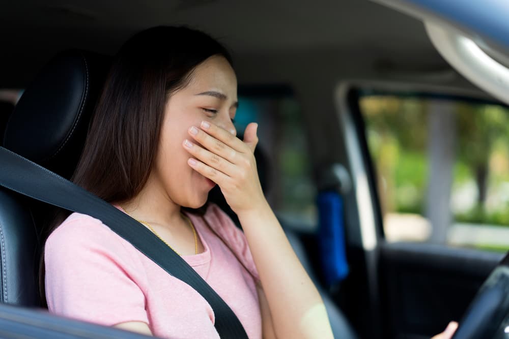 Portrait of an Asian female driver yawning from exhaustion while driving a car, highlighting the dangers of drowsy driving and the risk of traffic accidents.