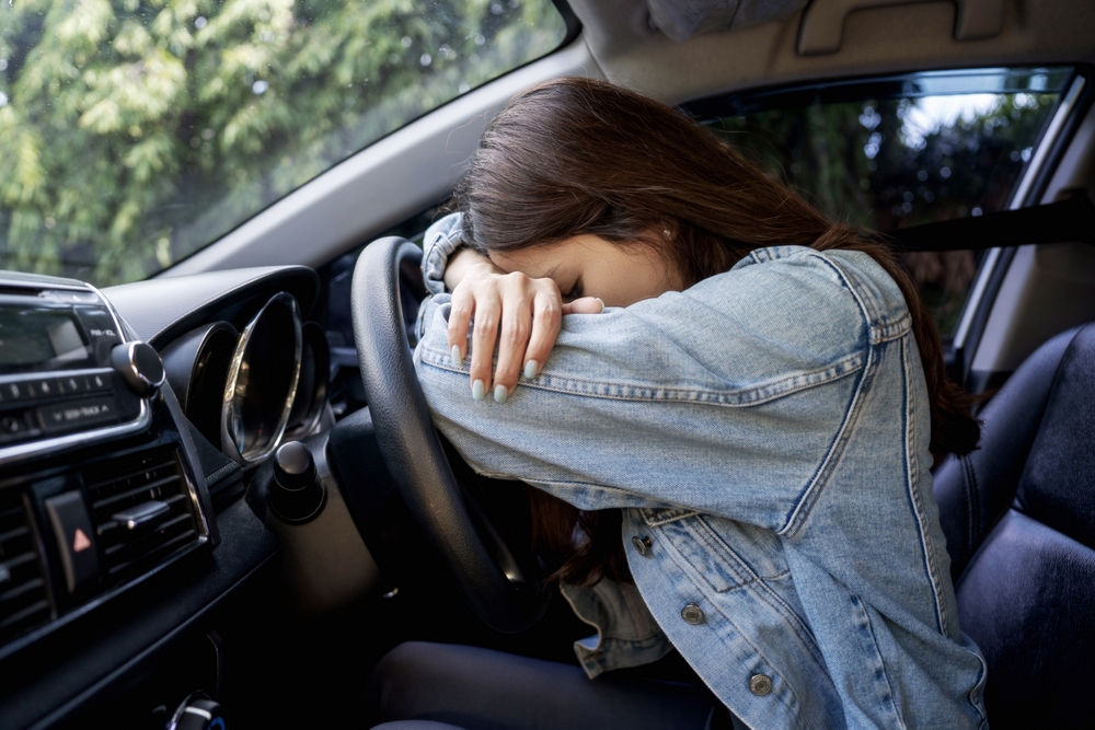 Asian girl looking stressed or exhausted, resting her head on the steering wheel inside a car.







