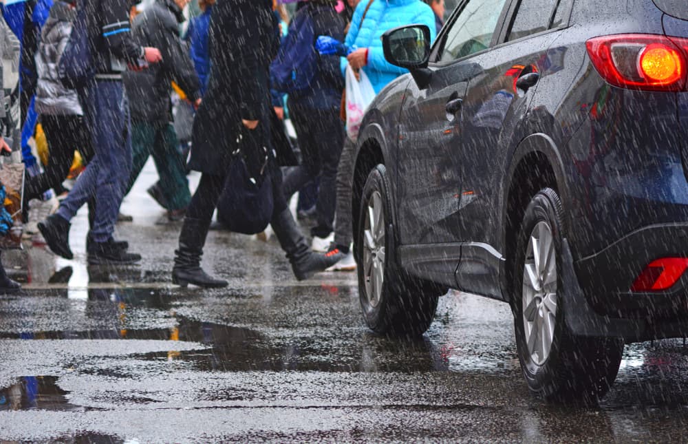 A crowd of people gathers on a pedestrian crosswalk beside a stopped car in a rainy city, emphasizing urban life and the challenges of navigating wet weather conditions.